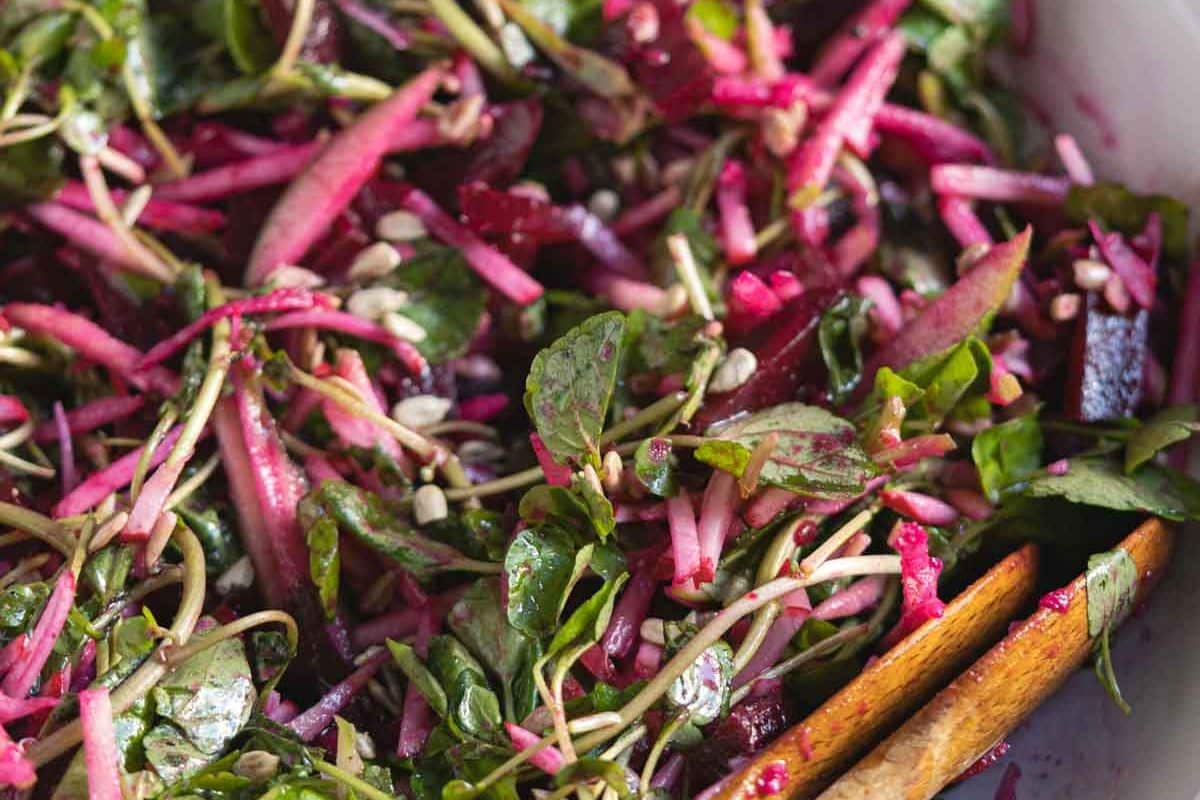Close-up of a fresh salad with leafy greens, shredded beets, seeds, and wooden salad tongs, showing vibrant pink and green colors—perfect for pairing with your favorite apple dishes during apple season.