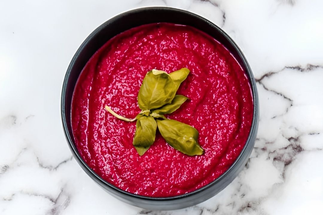 A bowl of bright pink beet hummus garnished with fresh basil leaves, placed on a white marble surface.