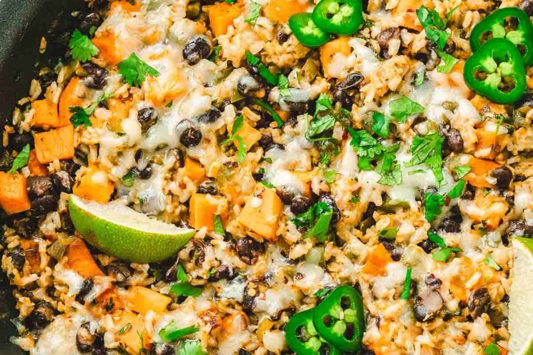 A close-up of a skillet dish with rice, black beans, sweet potatoes, melted cheese, jalapeño slices, fresh cilantro, and lime wedges.