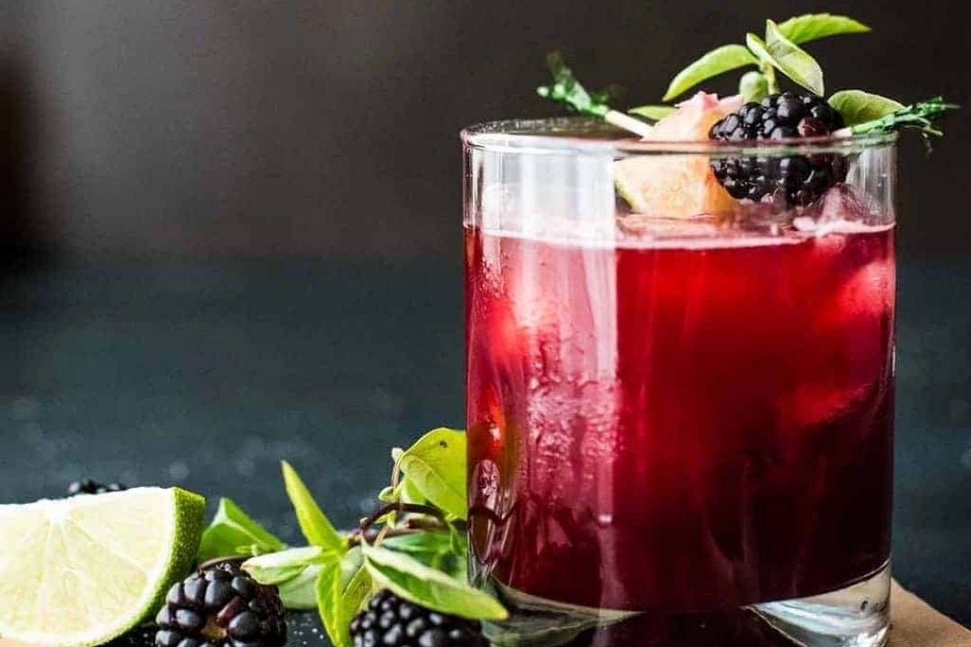 A glass of dark red blackberry cocktail, perfect for sipping, is garnished with blackberries, lime, and herbs as it sits on a dark surface.