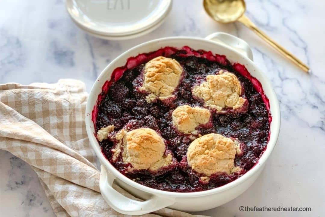Round white dish with baked berry cobbler, topped with five golden-brown biscuits, on a checkered cloth with a gold spoon nearby—a perfect choice for easy desserts or delightful cookout desserts.