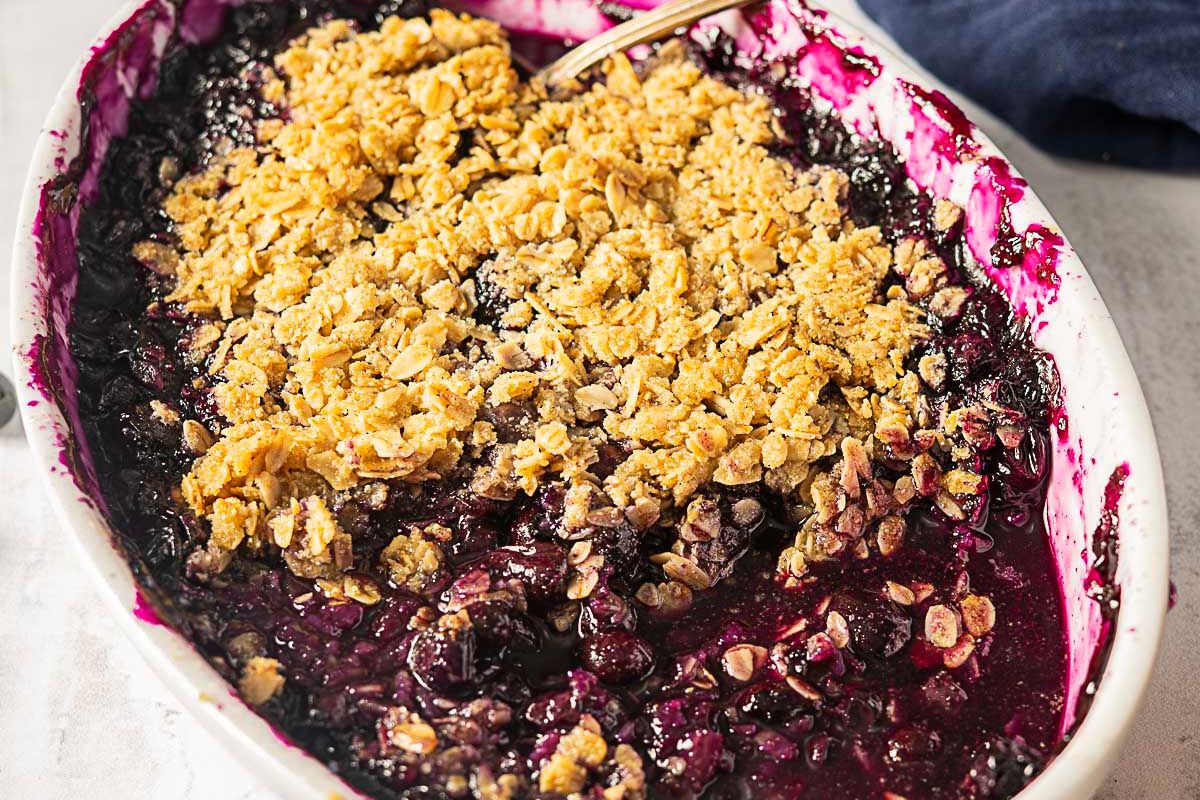 A baked berry crumble dessert in a white oval dish topped with a golden oat and crumble layer, with a serving spoon visible—perfect for those seeking delicious gluten-free recipes to enjoy at your next gathering.