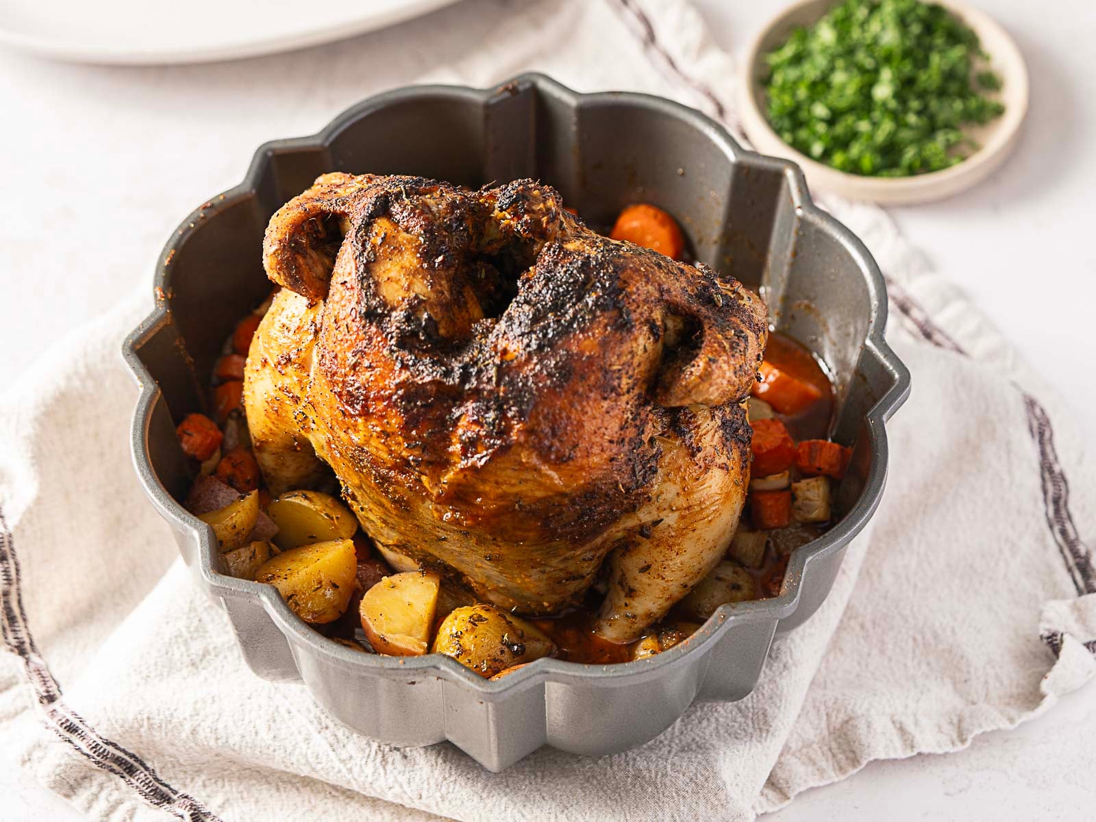 A roasted whole chicken, inspired by inventive new chicken dishes, sits in a bundt pan with potatoes and carrots, placed on a white towel. A small dish of chopped herbs is in the background.