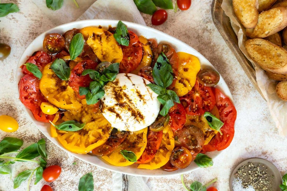 An oval plate of sliced yellow and red tomatoes, fresh basil, and burrata cheese drizzled with balsamic glaze, next to bread slices and a bowl of pepper—perfect for showcasing the vibrant flavors of summer recipes.