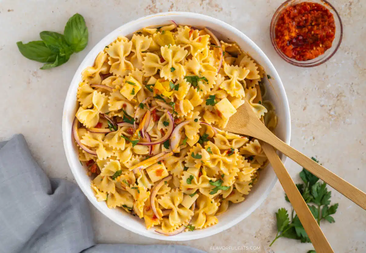 Bowl of bowtie pasta salad with herbs, red onions, and cheese, next to a jar of chili oil, basil, parsley, and a napkin. Wooden salad servers rest in the bowl, making it a perfect make-ahead option for stress-free hosting and ideal for your Labor Day sides spread.