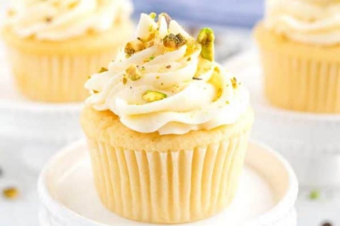 Vanilla cupcake with swirled white frosting, topped with chopped pistachios, displayed on a white plate with two similar cupcakes in the background.