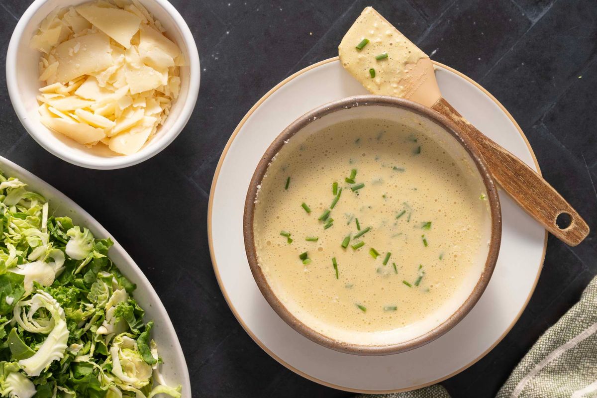 A bowl of creamy soup garnished with chopped chives sits on a plate, next to a bowl of shaved cheese and a dish of sliced greens—perfect accompaniments for enjoying with the best dressings or your favorite homemade dressings.