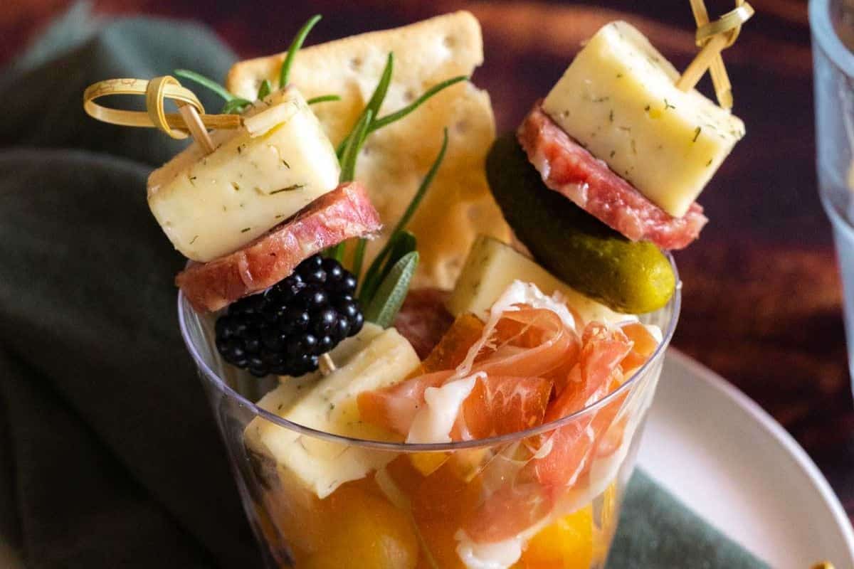 Glass contains skewers with cheese, salami, pickles, and a blackberry, alongside slices of prosciutto, cheese cubes, yellow tomatoes, and a cracker—perfect for those looking for fresh recipes and creative entertaining ideas.