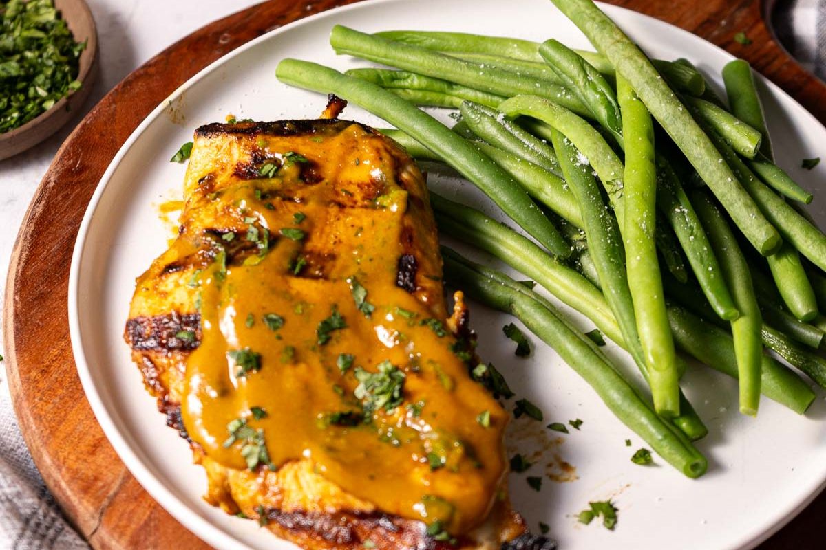 Grilled chicken breast topped with yellow sauce and chopped herbs, served with a side of steamed green beans on a white plate—perfect for your collection of flavorful summer recipes.