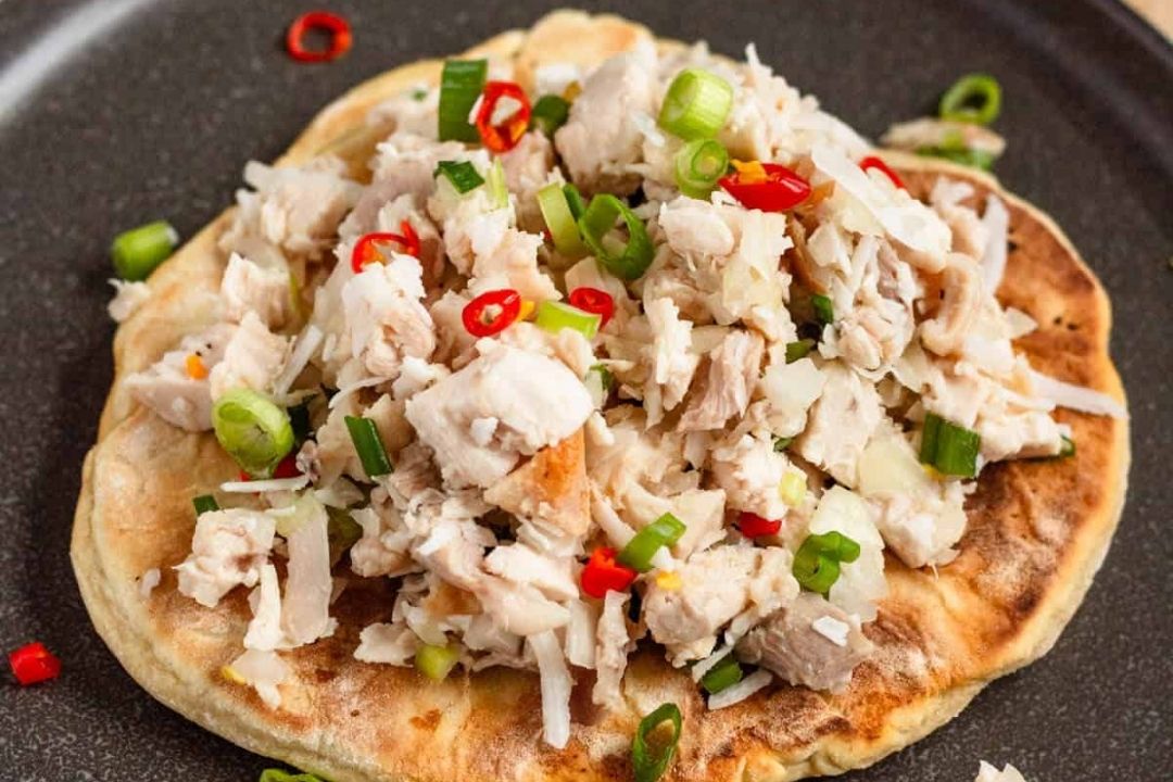 A flatbread topped with chopped cooked chicken, sliced green onions, red chili slices, and shredded white vegetables on a dark plate.