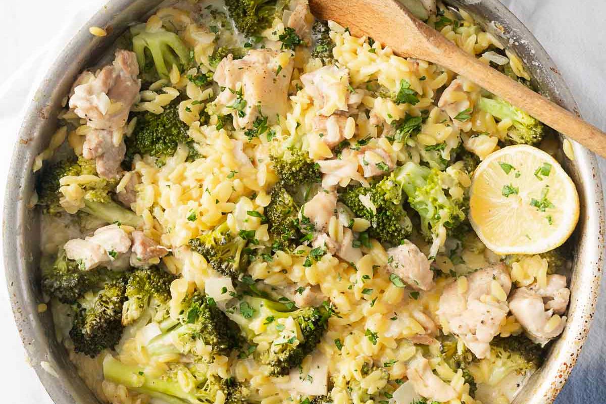 A skillet with cooked orzo pasta, pieces of chicken, broccoli florets, a lemon slice, and a wooden spoon, garnished with chopped herbs—perfect for cozy pasta dinners and inspiring new dinner ideas.