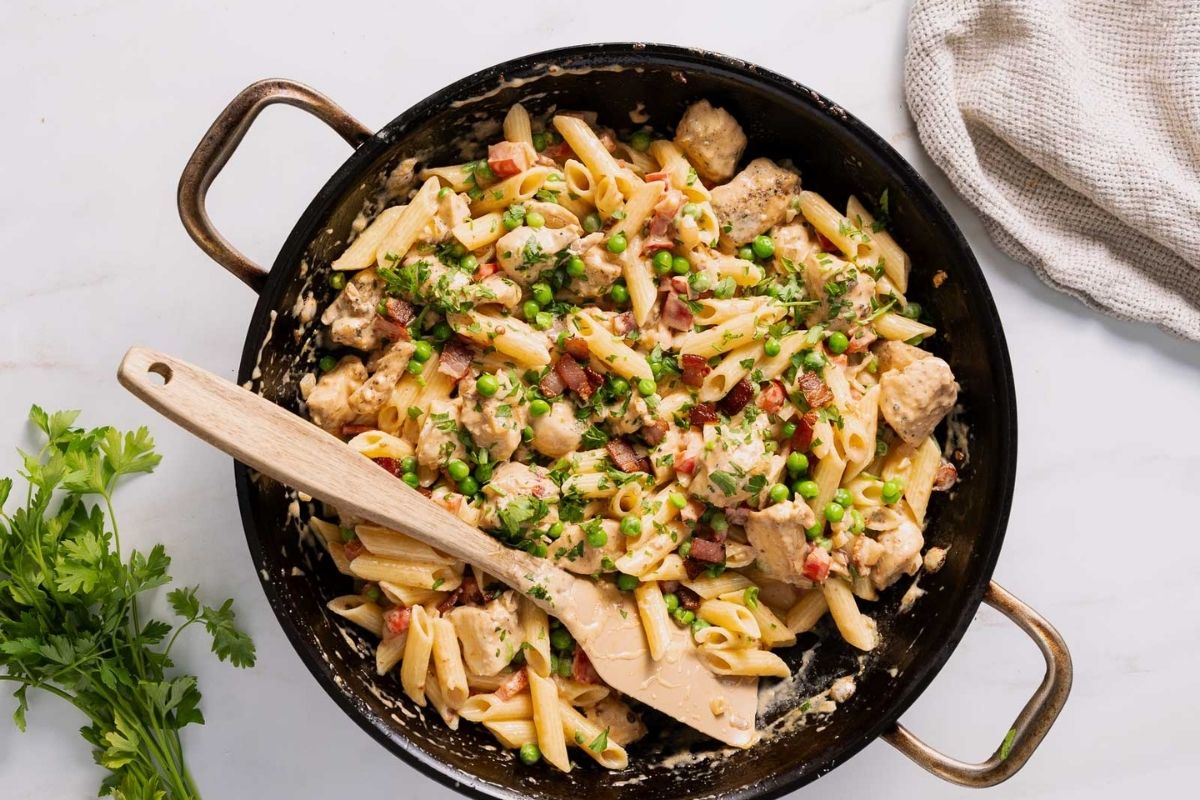 A skillet filled with penne pasta, chicken, peas, bacon, and creamy sauce, topped with chopped herbs; a wooden spoon rests inside the pan—perfect inspiration for cozy pasta dinners and new dinner ideas.