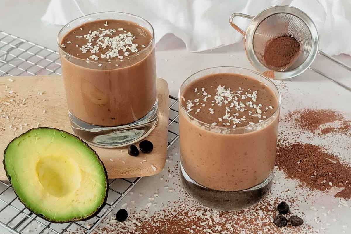 Two glasses of chocolate avocado smoothie topped with shredded coconut, surrounded by cocoa powder, an avocado half, and a metal sieve on a cooling rack—an irresistible treat for chocolate lovers searching for new chocolate recipes.