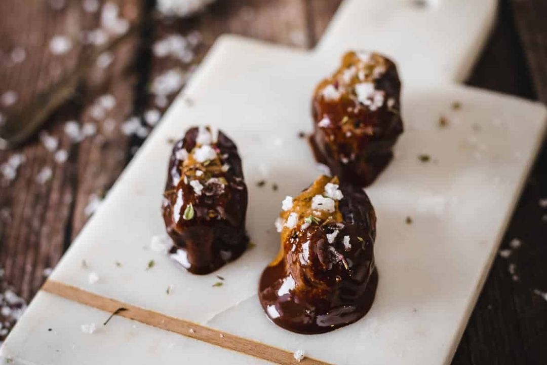 Three chocolate-covered dates topped with sea salt sit on a white marble cutting board with a wooden edge, placed on a rustic wooden surface—an irresistible treat for chocolate lovers and perfect inspiration for your favorite chocolate desserts.