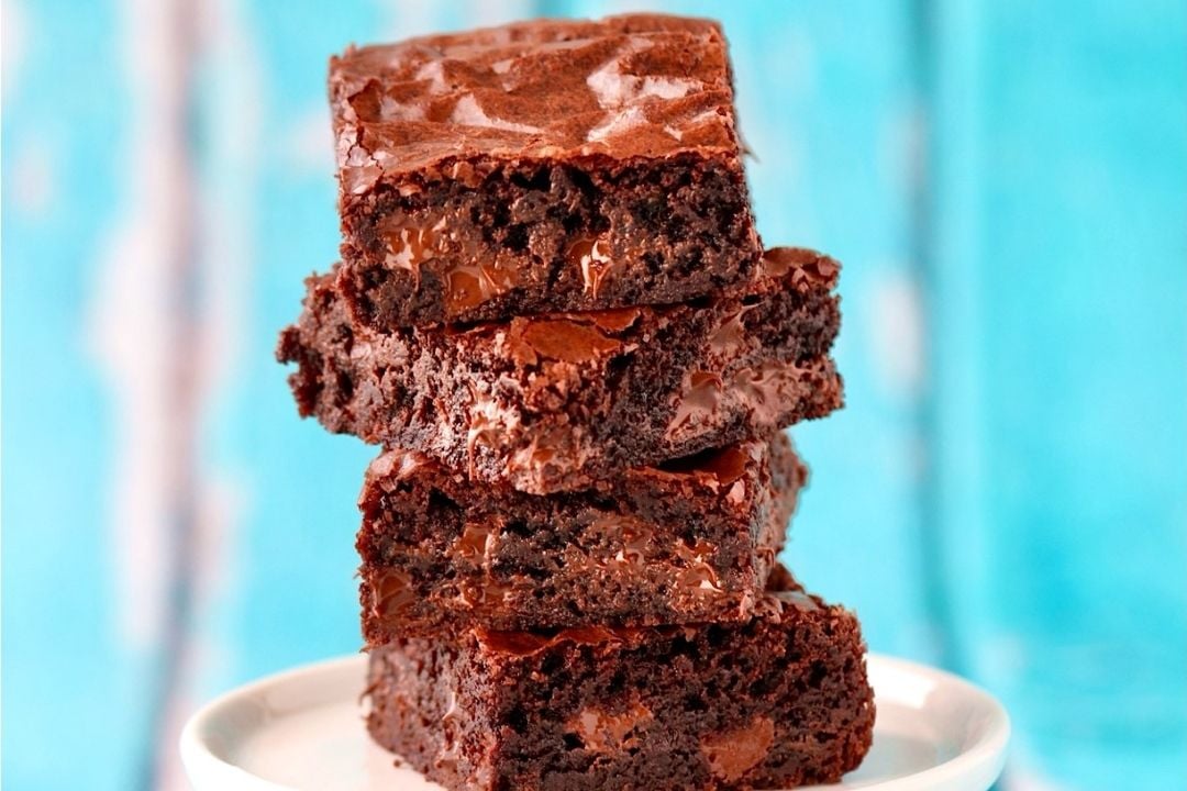 A stack of four chocolate brownies with a shiny, crackled top is placed on a white plate against a blue background, making these rich treats perfect for any chocolate lover.