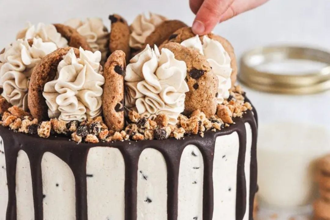 A hand places a chocolate chip cookie on top of a cake decorated with cookie treats, frosting swirls, and chocolate drip, with milk in the background.