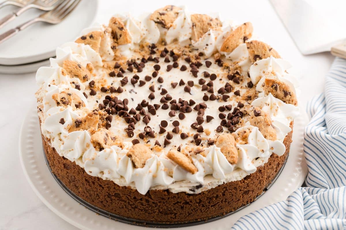 A round cookie cake with whipped cream, chocolate chips, and cookie pieces on top—reminiscent of classic chocolate chip cookies—is displayed on a white plate next to a blue-striped napkin.