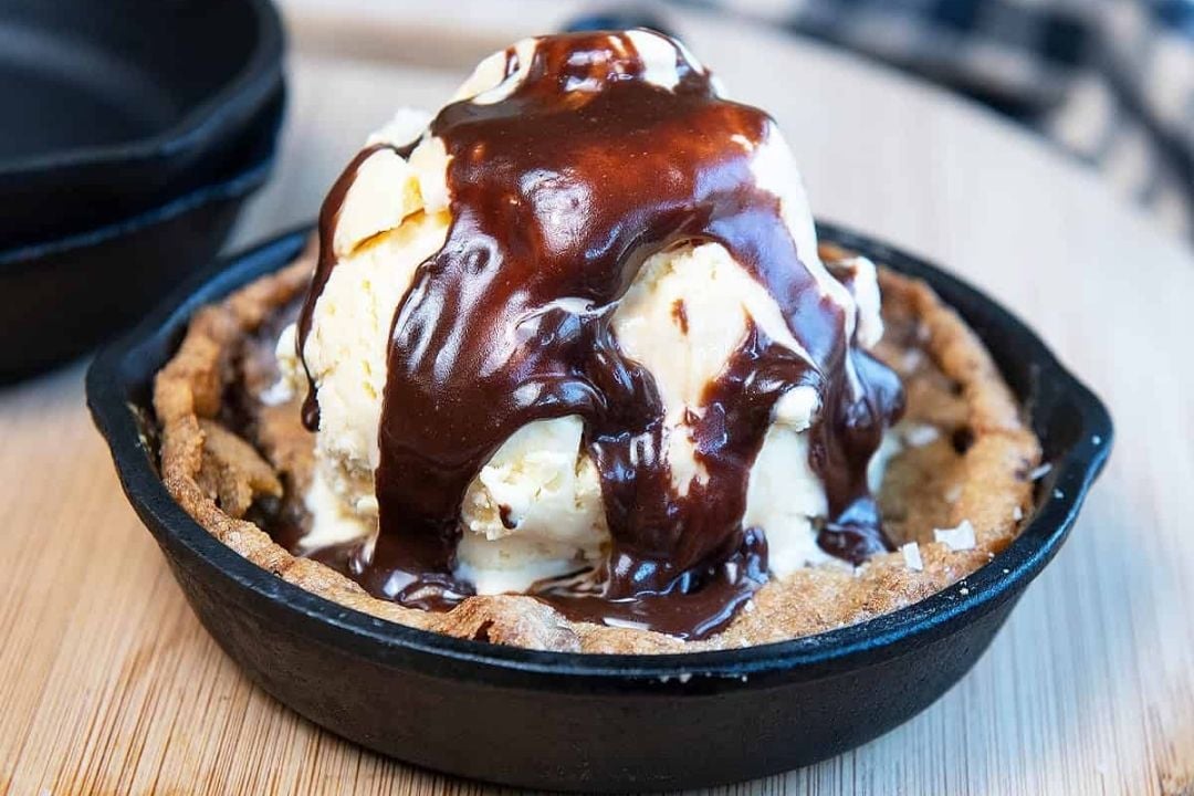 A scoop of vanilla ice cream sits on a chocolate chip cookie in a small skillet, topped with chocolate sauce, on a wooden surface—a delicious twist on classic chocolate chip cookie treats.