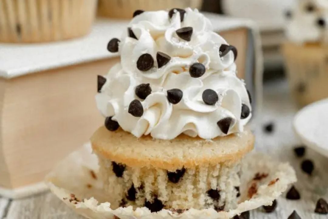 A chocolate chip cupcake with whipped cream and chocolate chips on top, sitting on an unwrapped cupcake liner, offers all the deliciousness of classic cookie treats in a decadent, bite-sized form.