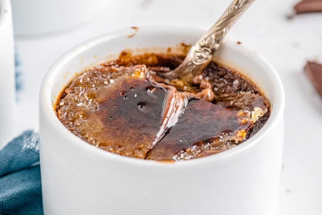 A white ramekin filled with chocolate creme brulee, featuring a caramelized sugar top and a silver spoon breaking through the surface—an irresistible treat perfect for recipes for chocolate lovers.