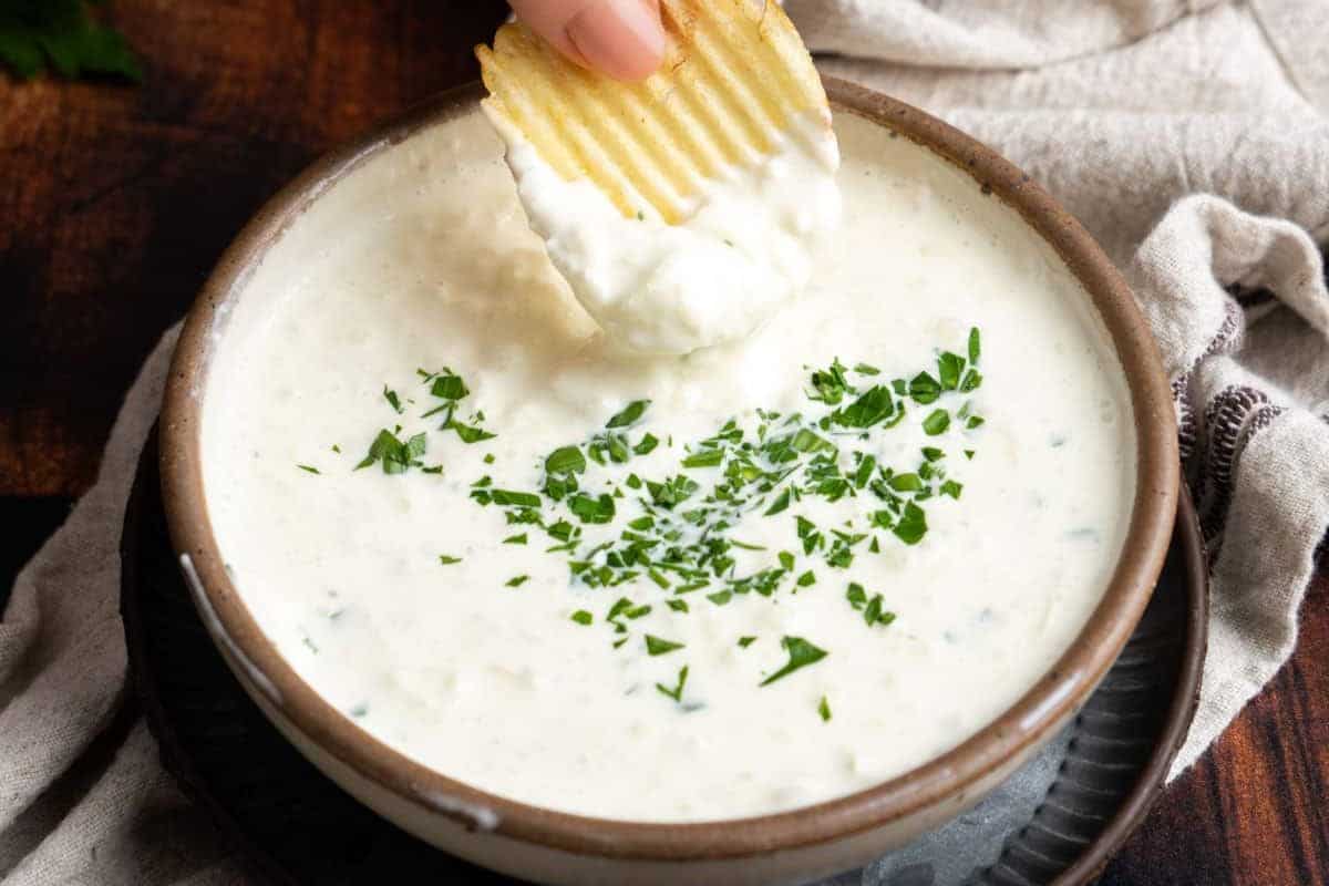 A hand dips a ridged potato chip into a bowl of creamy cold dip topped with chopped herbs, sitting on a dark plate beside a beige cloth—perfect for effortless snacking during game day gatherings.