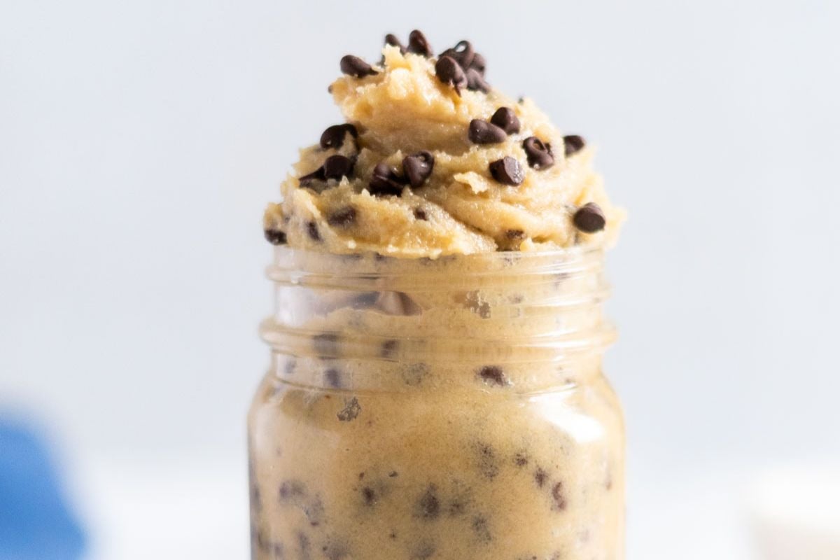 A glass jar filled with edible cookie dough topped with mini chocolate chips, set against a light background—perfect for anyone who loves chocolate chip cookie recipes and delightful cookie treats.