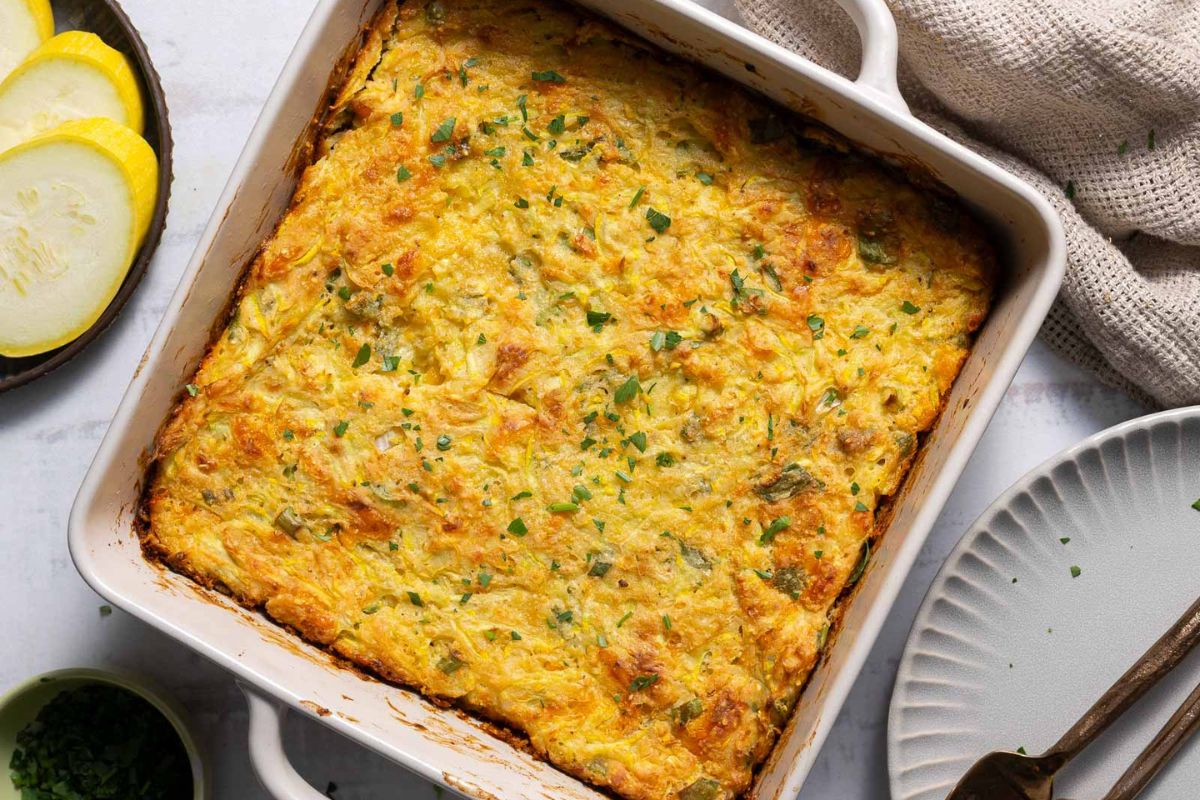 Baked yellow squash casserole in a square white dish, garnished with chopped herbs, next to a plate, fork, and sliced yellow squash—this classic is one of those tried-and-true recipes that always stands out among popular potluck dishes.