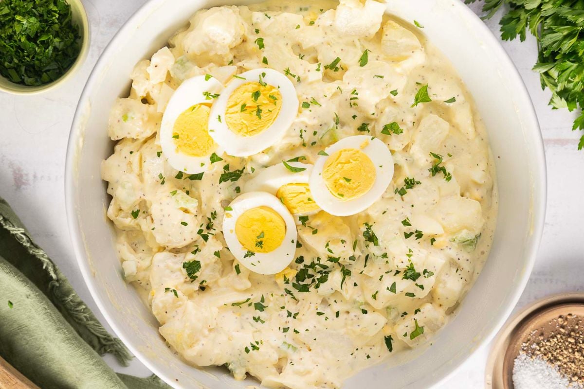 A bowl of creamy potato salad topped with sliced hard-boiled eggs and chopped parsley, surrounded by small bowls of ingredients and seasonings—perfect for adding to your collection of Labor Day cookout recipes.