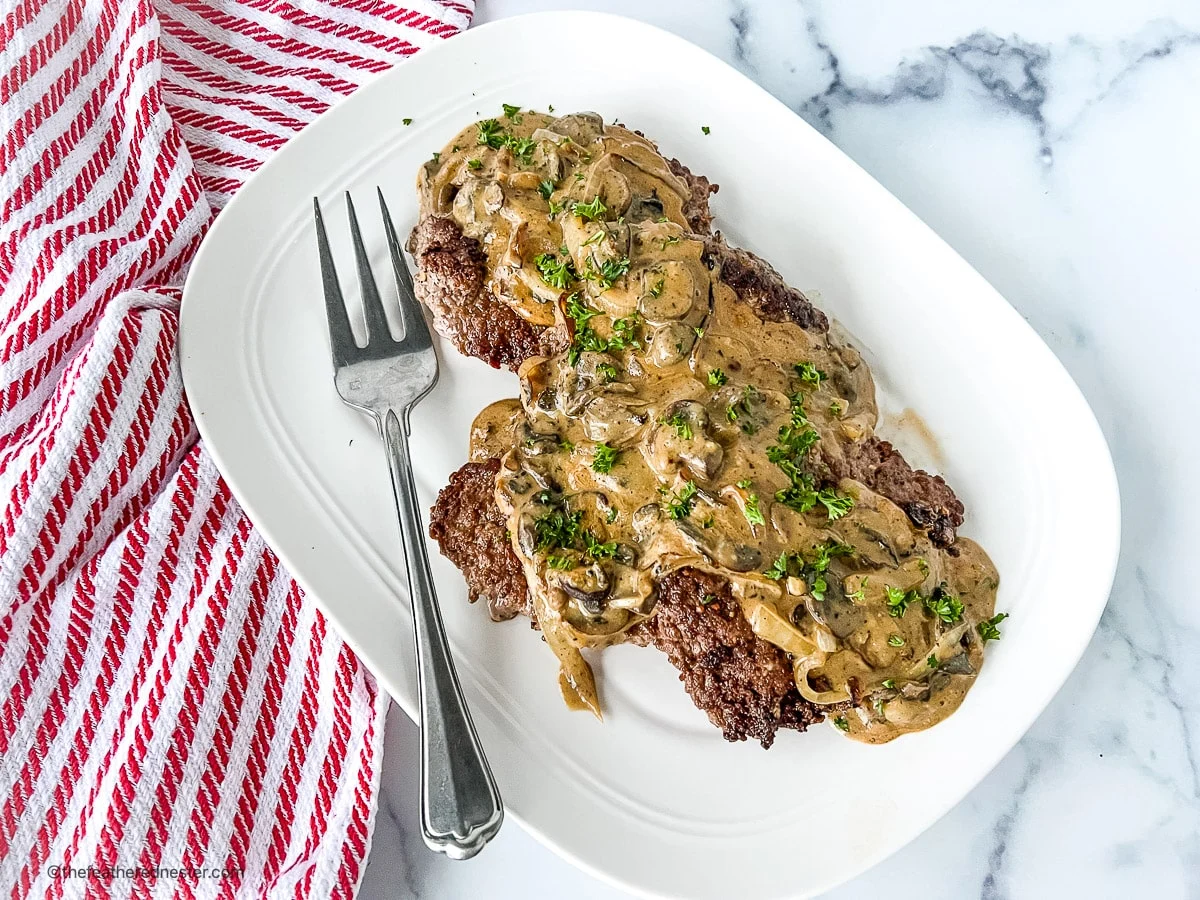 A plate of Salisbury steak topped with mushroom gravy, garnished with chopped parsley, sits next to a serving fork on a white platter with a red and white striped towel—an inviting dish perfect for cozy fall recipes or easy crockpot recipes.