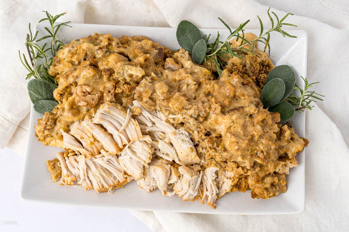 Rectangular white plate with shredded chicken breast covered in creamy gravy, garnished with fresh rosemary and sage sprigs, on a white cloth background—a comforting dish perfect for fall recipes and cozy Crockpot recipes.