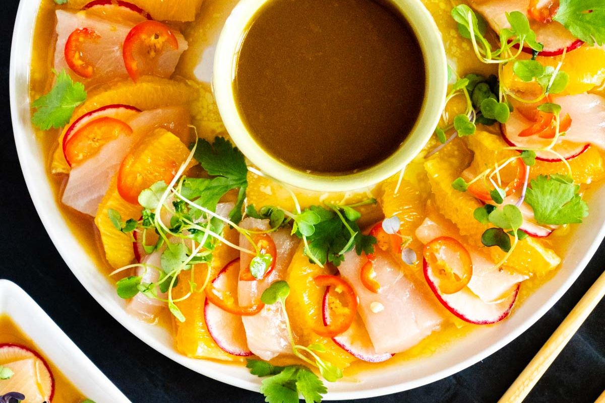 Sliced fish, orange segments, radishes, and greens arranged around a bowl of brown sauce on a white plate, making it perfect for guest meal ideas or anyone seeking fresh recipes.