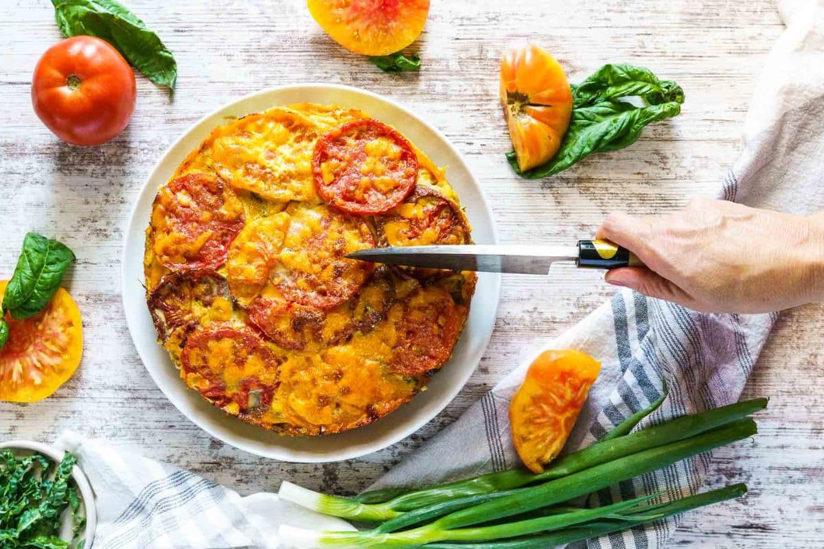 A hand slices a tomato and cheese casserole on a plate, surrounded by fresh tomatoes, basil leaves, green onions, and a striped cloth on a light wooden surface—perfect for those seeking delicious Garden-Fresh recipes.