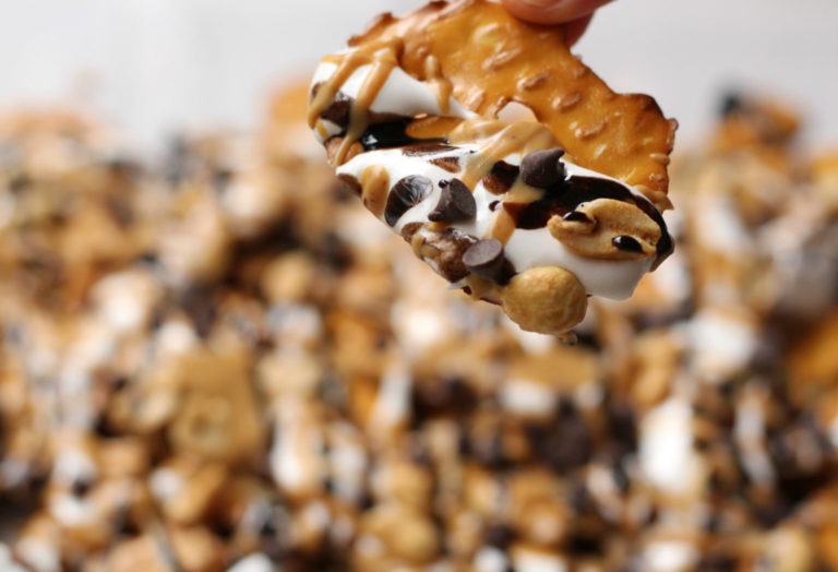 A hand holds up a pretzel covered with melted marshmallows, chocolate chips, and creamy peanut butter—a dreamy treat for fans of peanut butter desserts—with more of the same irresistible snack blurred in the background.