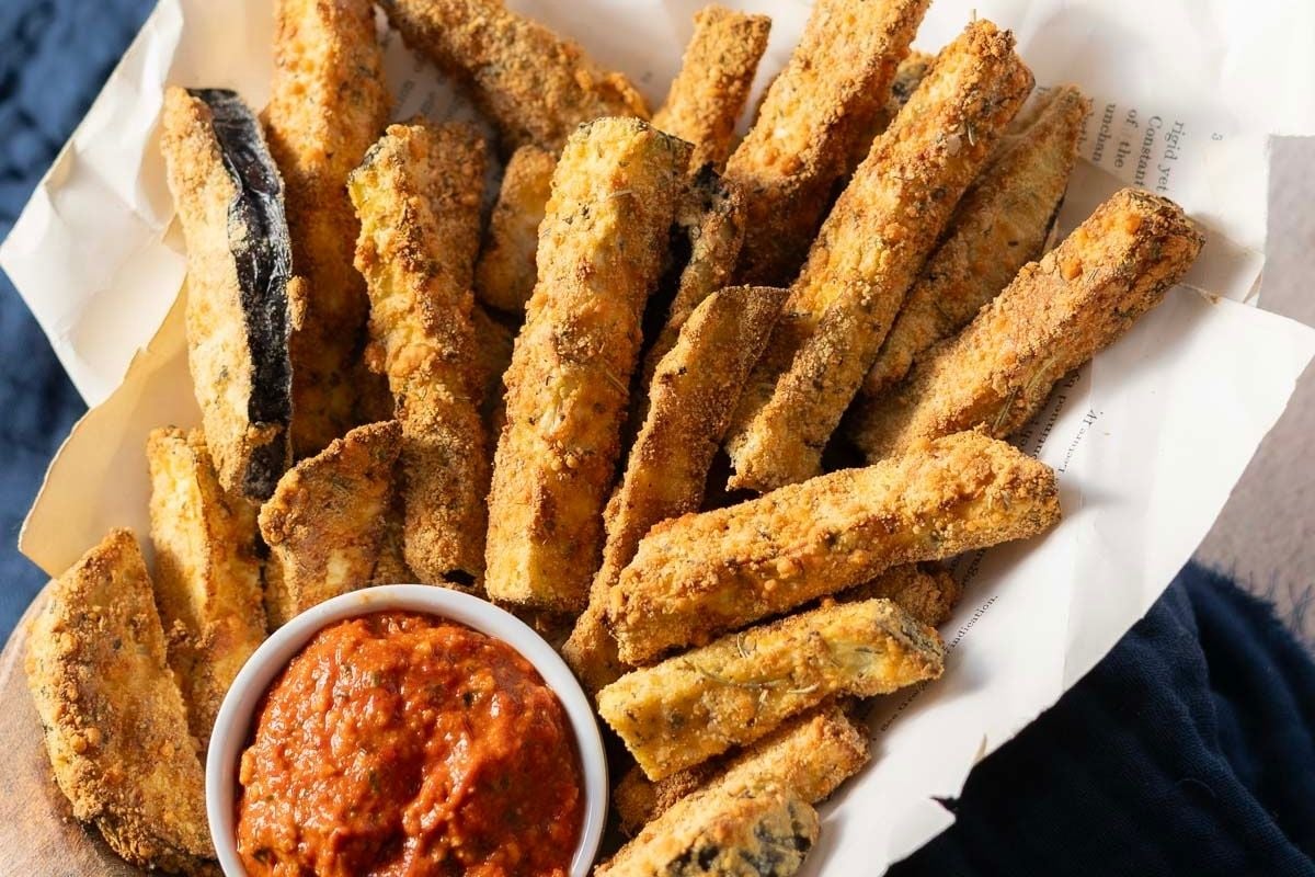 A pile of breaded zucchini fries on parchment paper is served with a small bowl of red marinara dipping sauce, making it one of the best easy vegetable recipes for a tasty snack or appetizer.