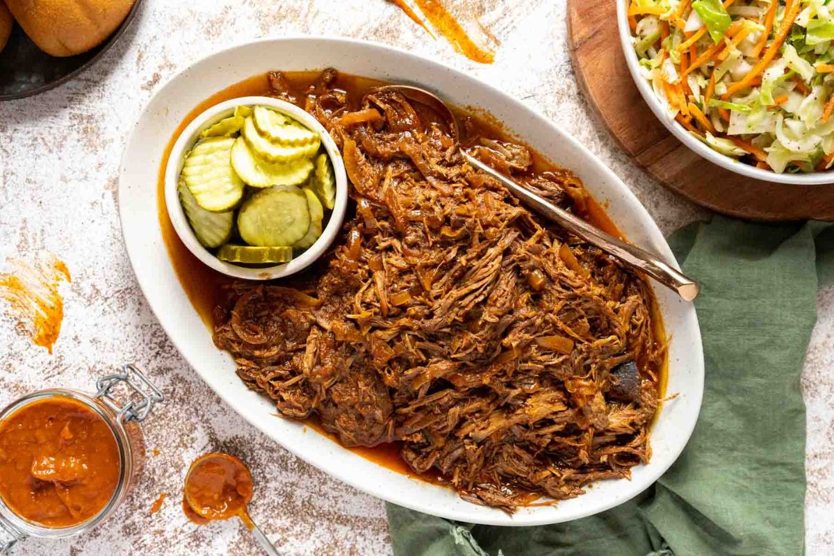 Oval platter of shredded barbecue beef with a bowl of sliced pickles, served next to a bowl of coleslaw and a jar of barbecue sauce—one of those tried-and-true recipes that never fails to be a crowd-pleaser at any gathering.