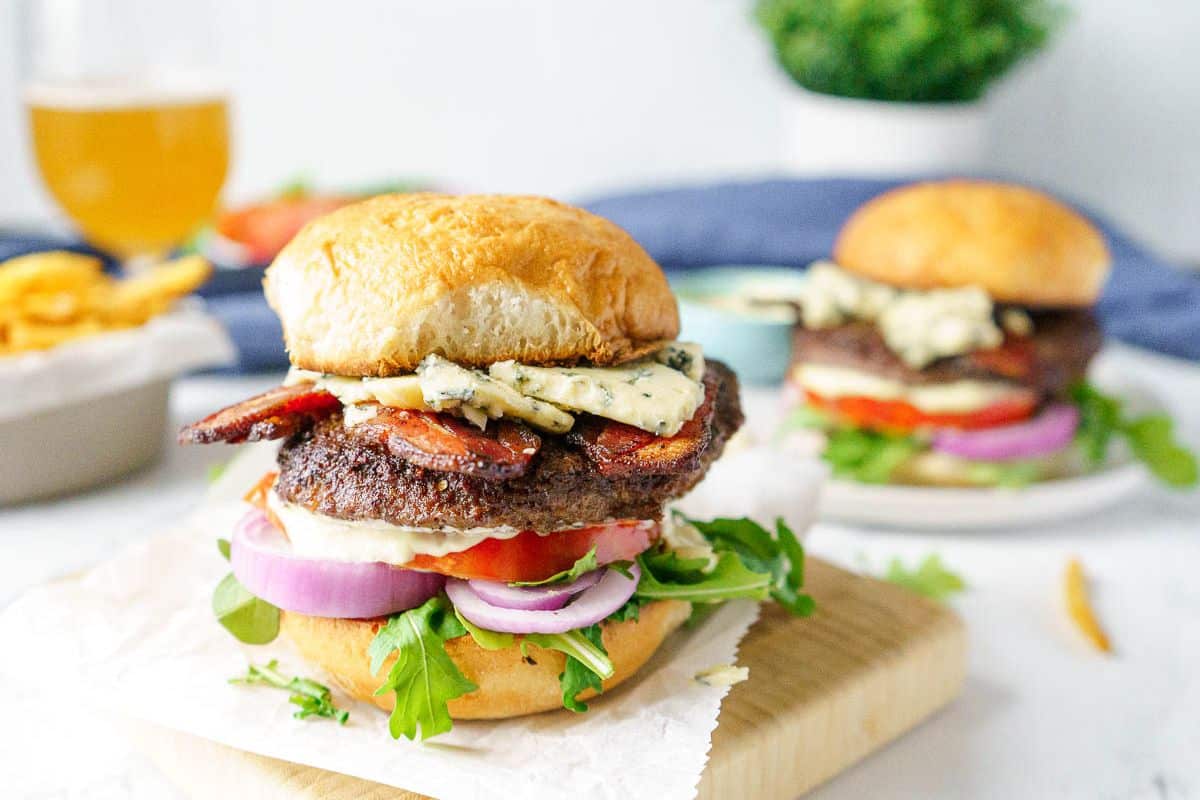 A hamburger with lettuce, tomato, red onion, bacon, and blue cheese crumbles on a bun sits on a wooden board, making it a mouthwatering addition to your Labor Day cookout recipes. Another similar burger is blurred in the background.