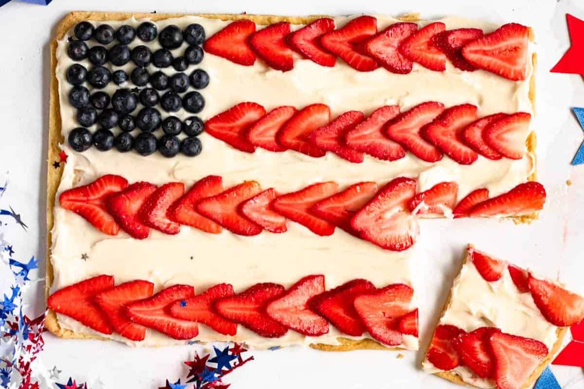 Rectangular fruit pizza decorated like a US flag with blueberries in one corner and rows of sliced strawberries on a cream-covered crust; one piece is cut out—a festive and easy dessert perfect for cookouts or Labor Day celebrations.
