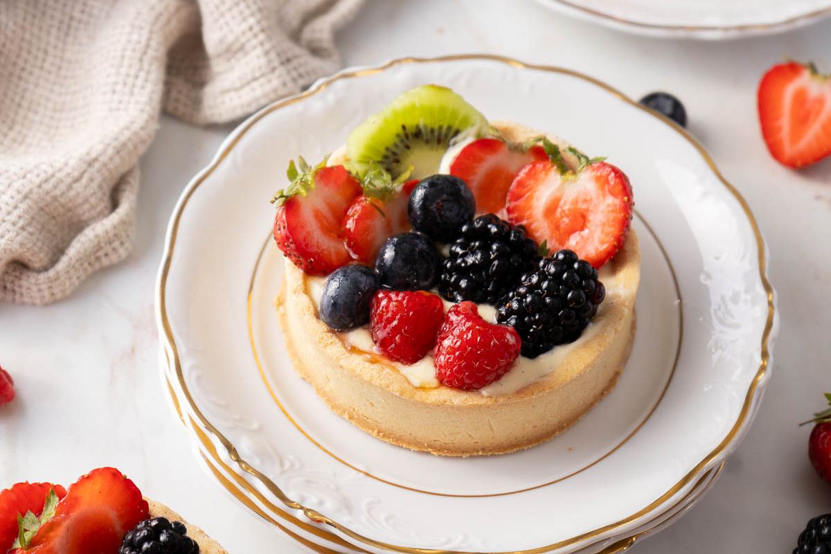 A fruit tart topped with strawberries, blackberries, blueberries, raspberries, and a slice of kiwi on a white plate with a gold rim—perfect for guest meals and ideal among fresh recipes.