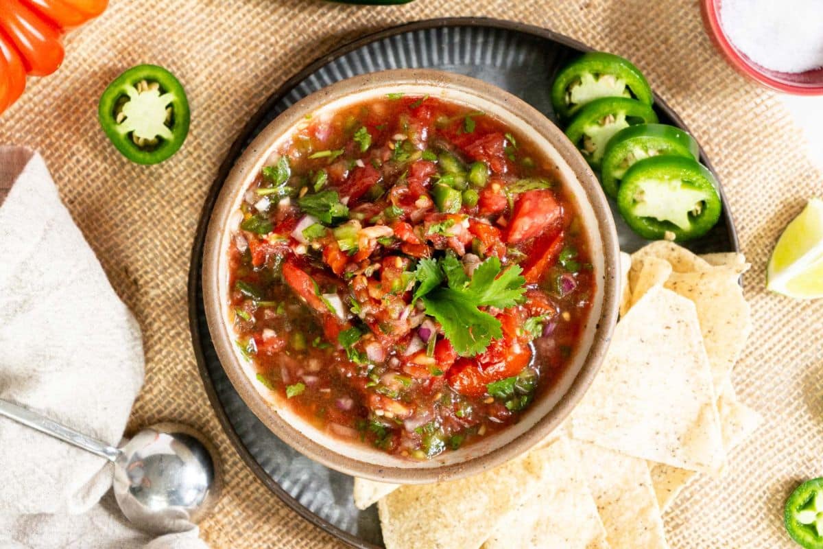 A bowl of fresh salsa with tomatoes, onions, and cilantro—perfect for garden tomato recipes—surrounded by tortilla chips, jalapeño slices, a lime wedge, and a metal spoon on a burlap surface.