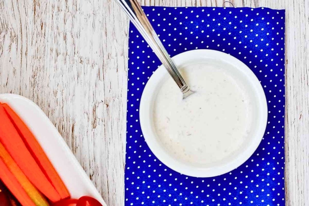 A bowl of ranch dressing with a spoon sits on a blue polka dot napkin beside a tray of sliced vegetables on a white wooden surface, showcasing one of the best dressings to elevate any fresh veggie platter.