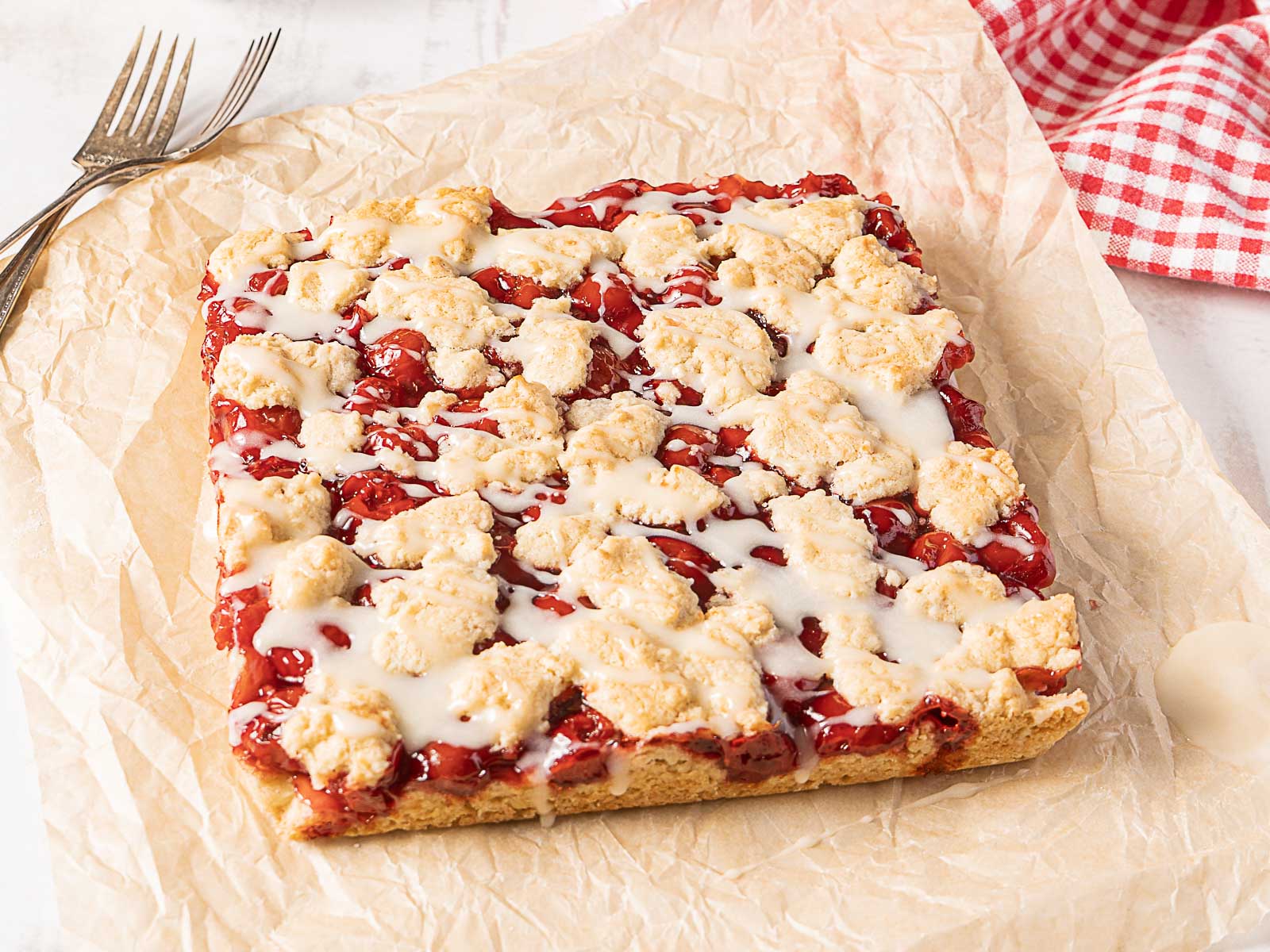 Square cherry crumble dessert with white icing drizzle on parchment paper, next to two forks and a red checked cloth—a delightful choice for cookouts or Labor Day gatherings, and one of the easiest desserts to share with friends.