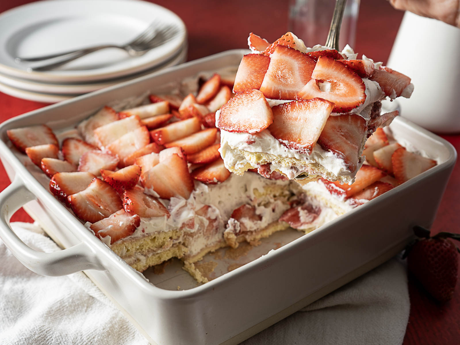 A square dish of strawberry shortcake with whipped cream and sliced strawberries, with one piece being lifted out, plates in the background—a delightful treat perfect for summer desserts and completely gluten free.