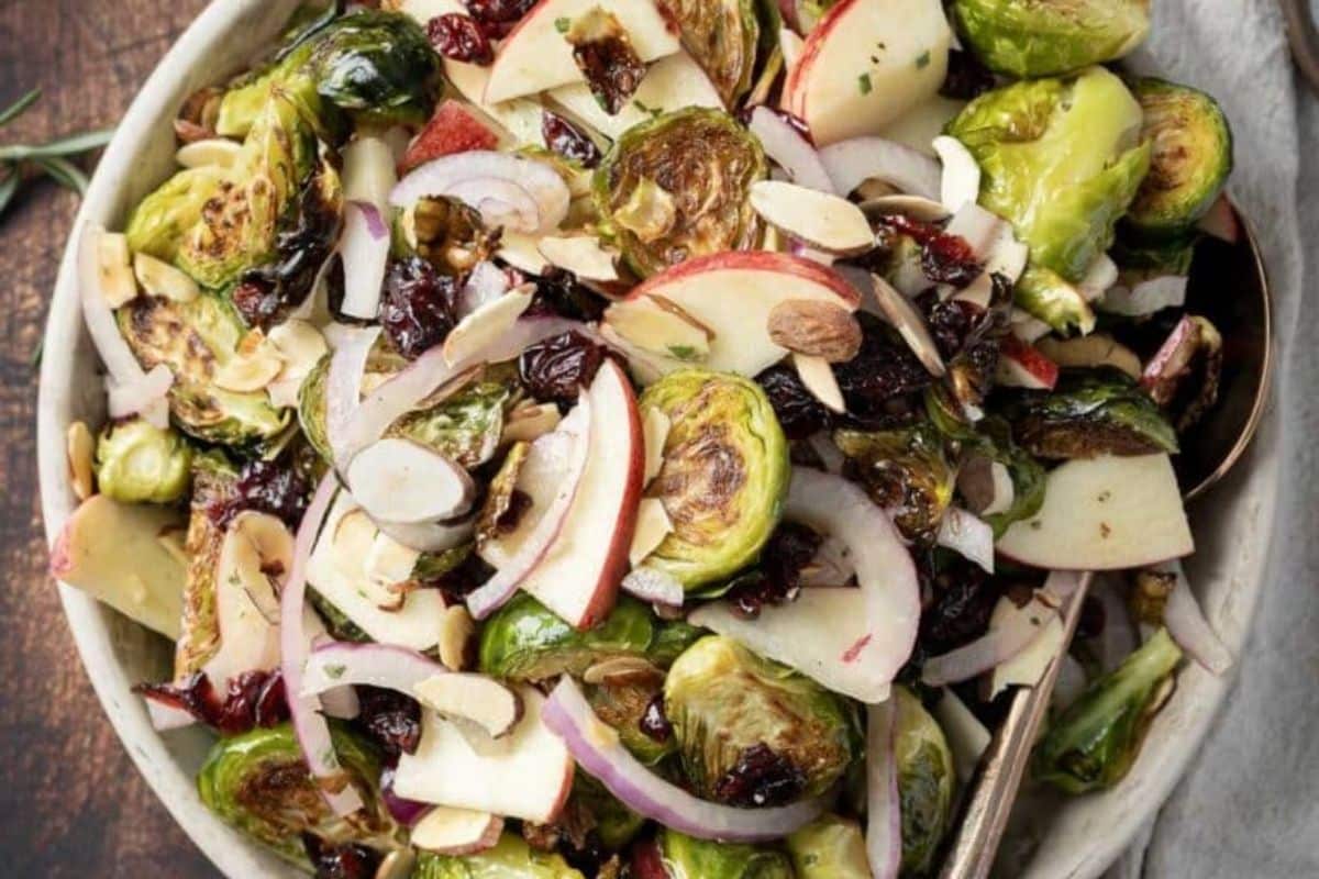 A bowl of salad with roasted Brussels sprouts, sliced apples, red onions, cranberries, and almond slivers, served with a metal spoon—one of our reader favorites and the perfect addition to your collection of popular side dishes.