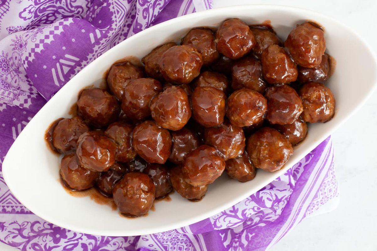 Oval white dish filled with glazed meatballs, placed on a purple and white patterned cloth—a perfect addition to your collection of crowd-pleasing recipes.