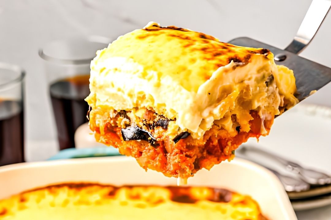 A spatula holds a slice of baked casserole with layers of creamy topping, vegetables, and tomato sauce over a baking dish. Two glasses and plates are in the background.