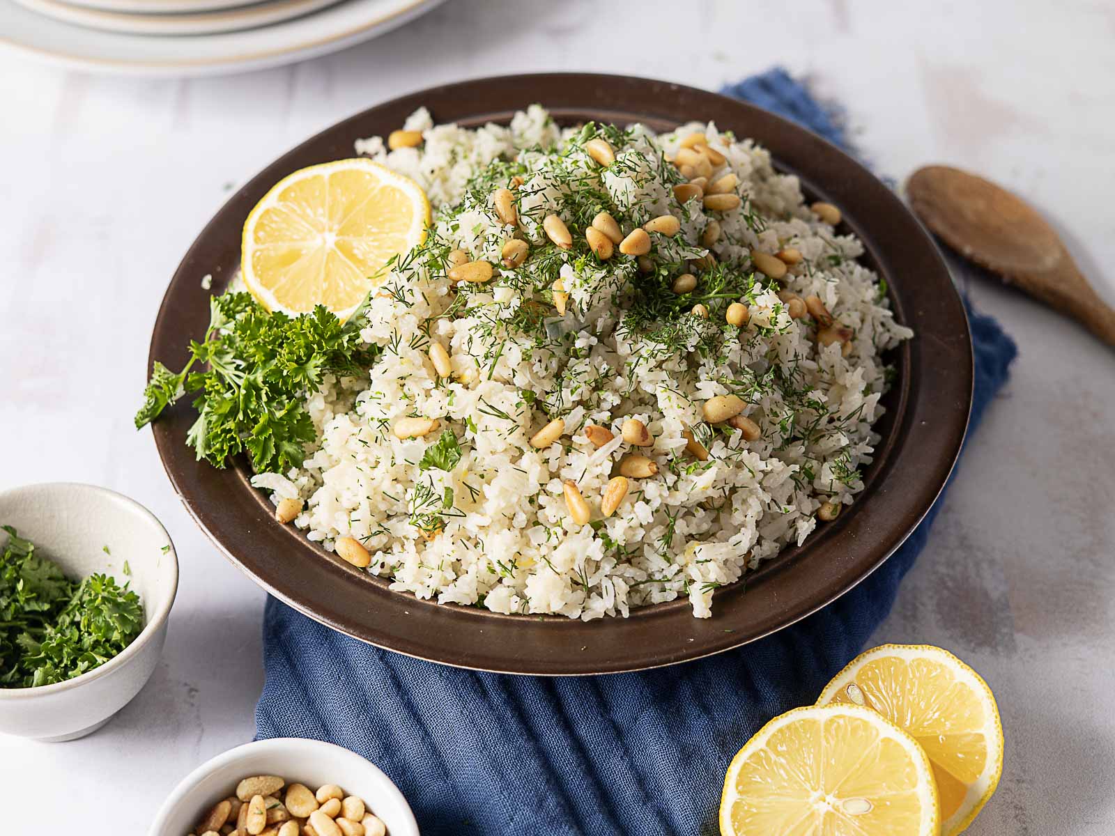 A plate of herbed rice garnished with pine nuts, fresh dill, parsley, and lemon slices, placed on a blue napkin with extra pine nuts and parsley nearby—perfect as one of those reader favorites that makes side dishes truly memorable.