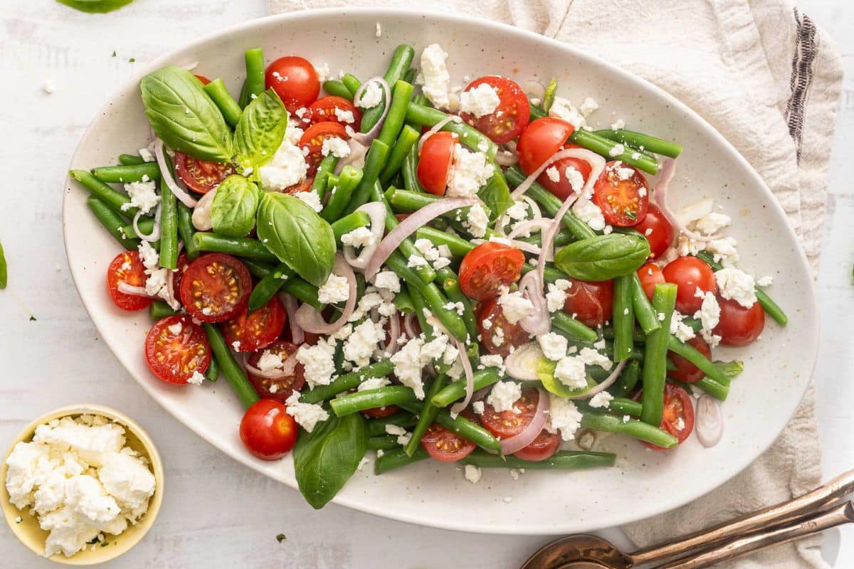 Oval platter with green beans, cherry tomatoes, sliced shallots, basil leaves, and crumbled feta cheese—an ideal Labor Day sides option for stress-free hosting. A small bowl of extra feta and serving utensils are nearby on a white cloth for easy, make-ahead serving.