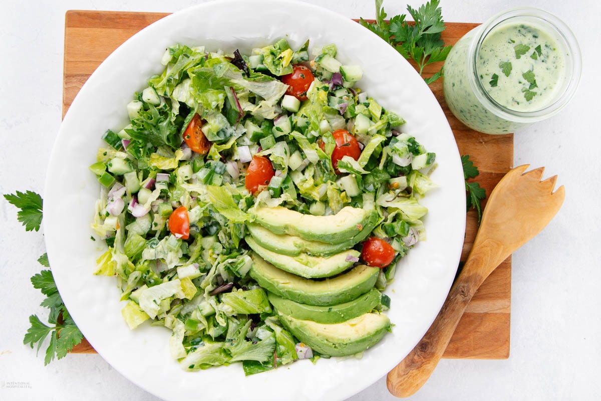 A bowl of chopped salad with lettuce, cucumber, tomato, red onion, and sliced avocado sits next to a jar of creamy green dressing and wooden salad utensils on a cutting board—a perfect example of Garden-Fresh summer recipes.
