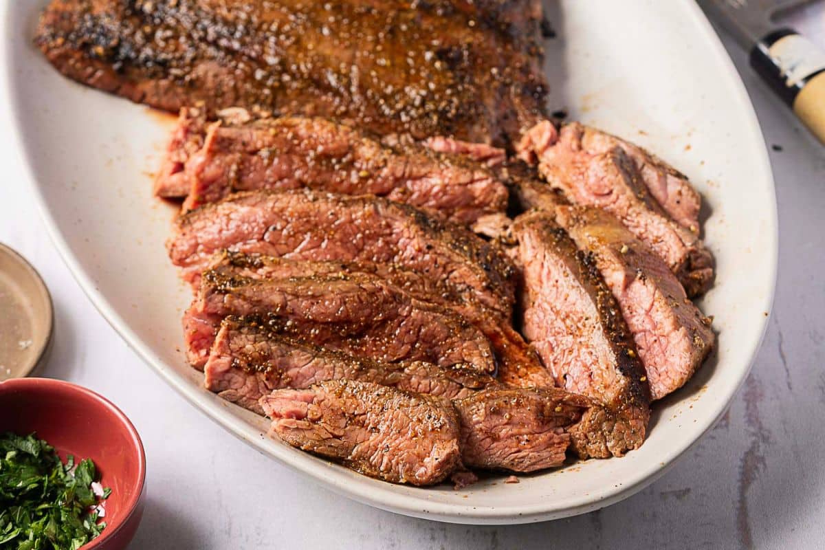 Sliced, seasoned steak on a white oval plate, showing a pink interior and a seared crust—perfect for gluten-free recipes or festive Labor Day recipes. Knife and herbs are visible nearby.