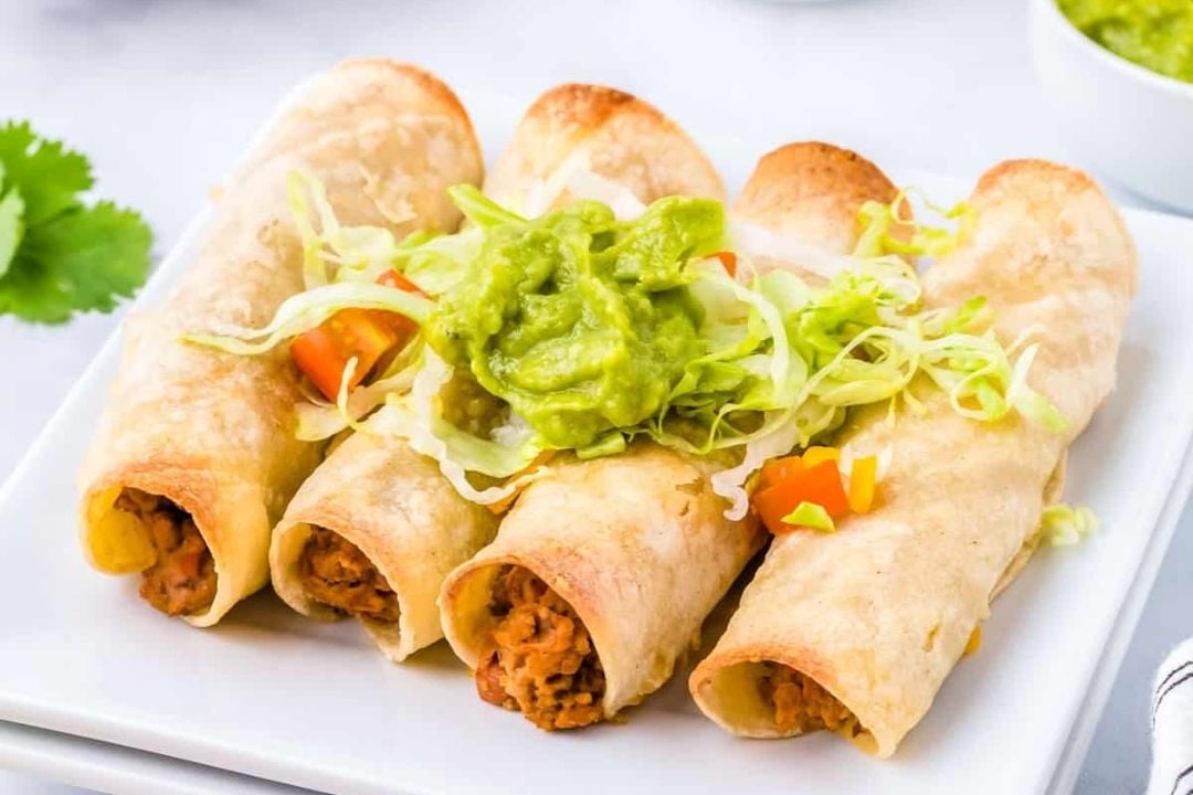 Four rolled tacos filled with meat, topped with shredded lettuce, diced tomatoes, and a scoop of guacamole, served on a white plate—perfect as one of your go-to Game Day Appetizers.